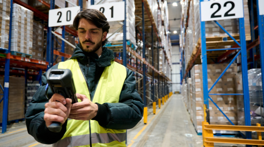 An adult male with dark hair and a beard scans inventory in a vast warehouse full of shelves, utilising a handheld scanner. He is wearing a dark jacket and bright yellow safety vest. The shelves are lined with boxes, and the warehouse stretches into the distance.